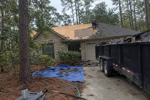 Roof tear-off in progress with bare decking and a debris trailer on a wooded lot