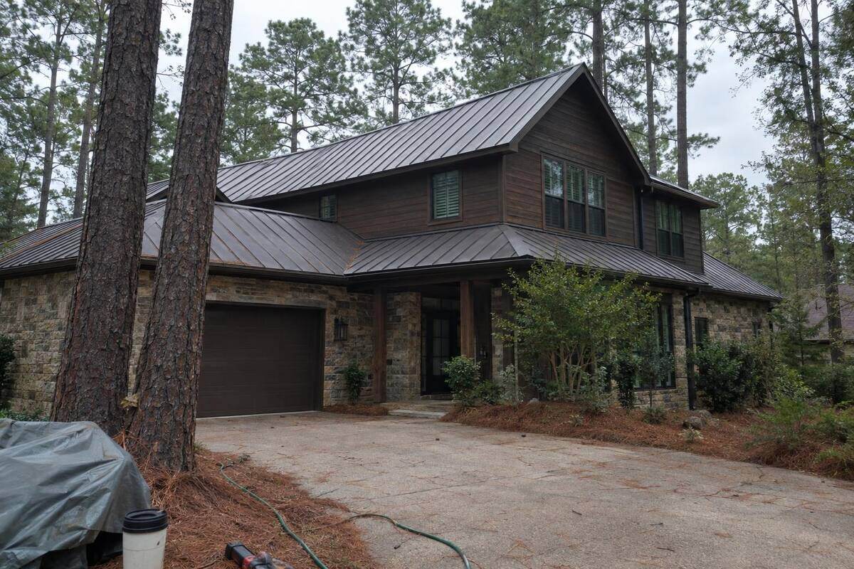 Dark bronze standing-seam metal roof on a two-story house with pine trunks in the foreground